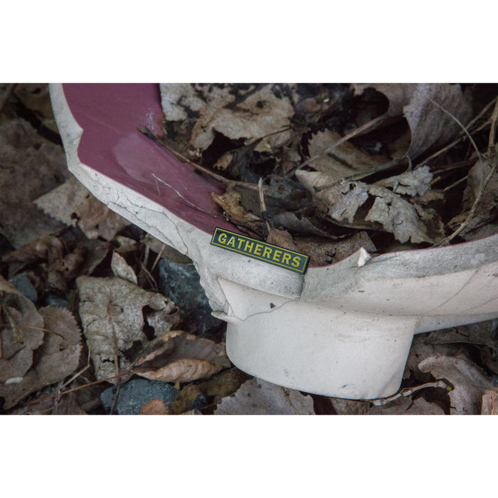 A broken white and red object lies among dry leaves, featuring a small yellow and black Gatherers labelβreminiscent of an Alive Beyond Repair enamel pin by Gatherers left behind.