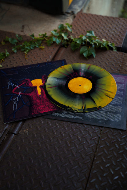 A yellow and black splattered vinyl record of Hail The Sun's Mind Rider rests on an album cover featuring abstract red and yellow art, placed on a metal surface with green ivy nearby.
