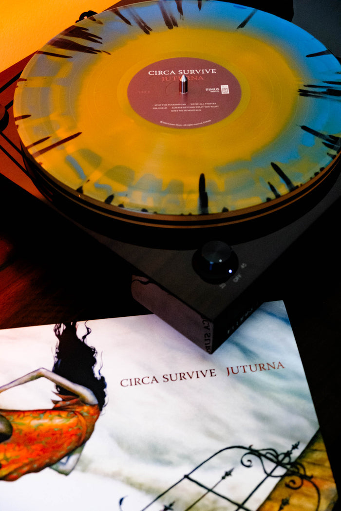 A splatter-patterned blue & orange starburst vinyl record plays on a turntable next to the album cover for Circa Survive’s Juturna, a limited edition LP which features surreal artwork of a woman in an orange dress falling.