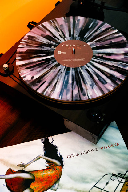 A black turntable plays a limited edition splattered LP record labeled Circa Survive Juturna. The album cover, showing an illustrated figure in an orange dress, lies nearby on a wooden surface under warm lighting.