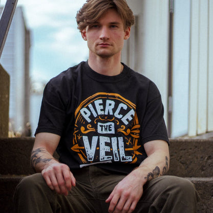 A young man with light brown hair and tattooed arms sits on concrete steps, wearing an olive green pants and a black Pierce The Veil T-shirt by Comfort Colors, looking seriously at the camera.