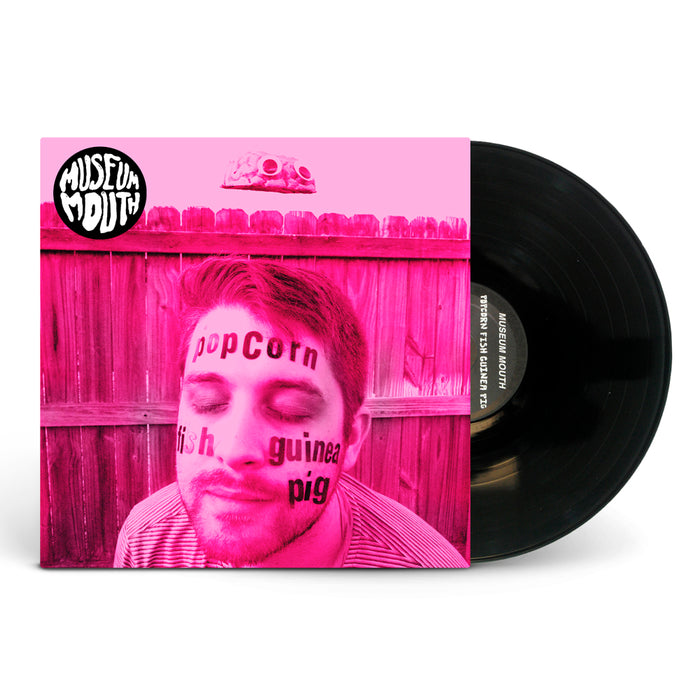 A vinyl record slides from its sleeve. The pink Museum Mouth album cover features a person with "popcorn," "guinea pig," and "fish" on their face, a wooden fence, and a brain illustration above them.