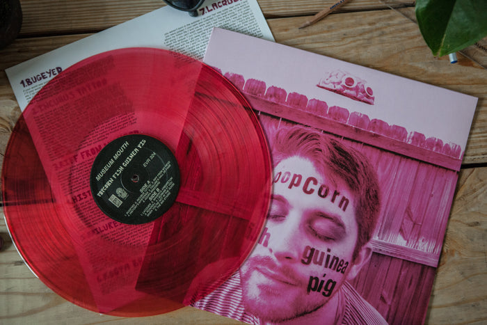 A Limited Pressing translucent red vinyl by Museum Mouth partially covers album art showing a man’s face with words like “popcorn” and “guinea pig”; set on wood, with a plant in the corner.