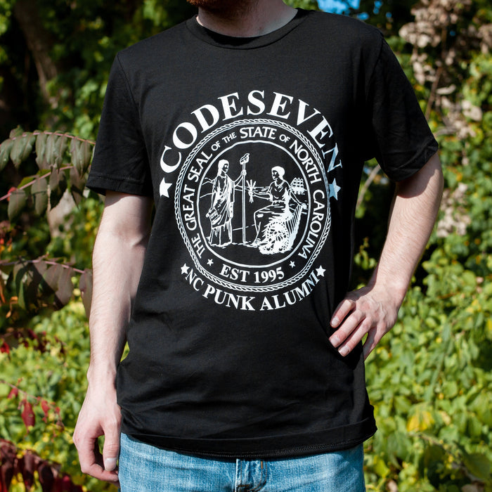 A model wearing a black T-shirt with a white design. The word "CODESEVEN" is surrounded by stars around the seal of the state of North Carolina. Within the seal it states 'THE GREAT SEAL OF THE STATE OF NORTH CAROLINA" and "EST. 1995." Below the seal are the words "NC PUNK ALUMNI" surrounded in stars. The model is pictured against a background of trees.