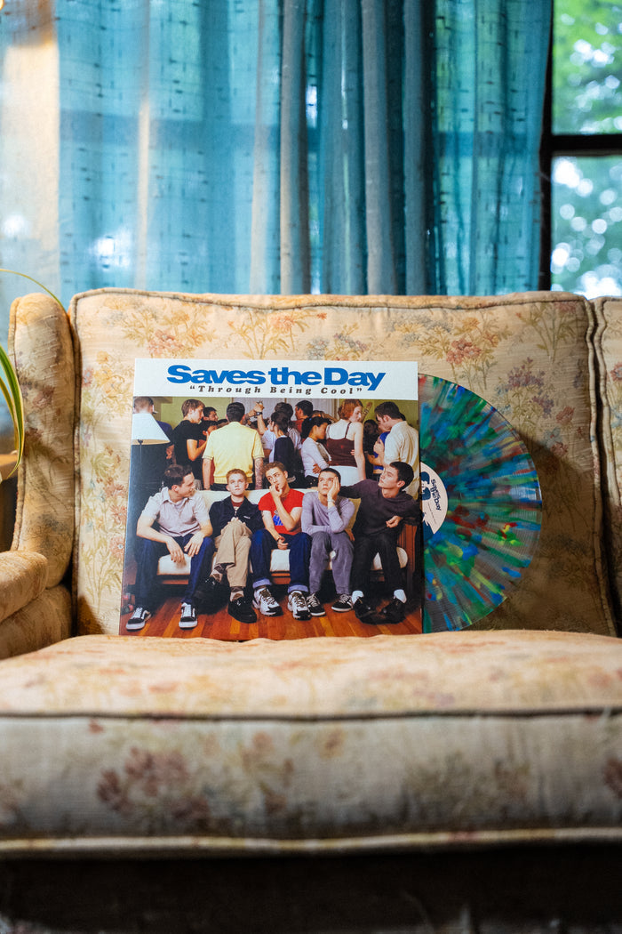 A Saves The Day vinyl record with a multicolored disc sits on a patterned beige couch, set in front of blue curtains and a window revealing greenery outside.