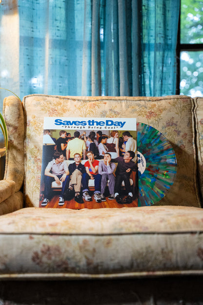 A Saves The Day vinyl record with a multicolored disc sits on a patterned beige couch, set in front of blue curtains and a window revealing greenery outside.