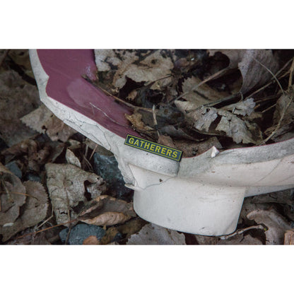 A broken white and red object lies among dry leaves, featuring a small yellow and black Gatherers label—reminiscent of an Alive Beyond Repair enamel pin by Gatherers left behind.